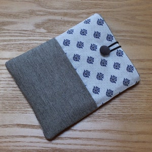 Puede incluir: Una funda para tablet de tela gris y blanca con un diseño estampado azul y blanco. La funda tiene un botón negro y un cierre elástico.