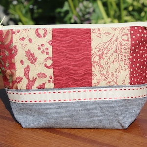 Könnte beinhalten: Eine rote und weiße Reißverschlusstasche mit einem grauen Stoffboden. Die Tasche besteht aus Patchwork-Stoff mit einem Blumen- und Blattdesign. Der Reißverschluss ist cremefarben und der Reißverschluss-Zipper ist ein kleiner Metallring. Die Tasche liegt auf einer Holzoberfläche.