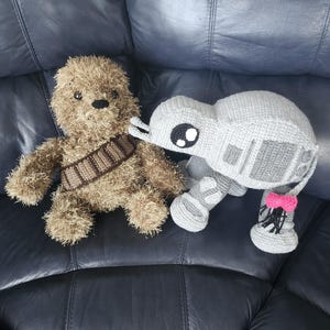 Op de afbeelding: Gehaakte pluche speelgoed van een bruin, harig Chewbacca-achtig personage met een bruine bandolier en een grijze AT-AT walker met een roze strik op zijn voet. Het speelgoed staat op een donkerblauwe leren ondergrond.