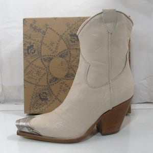 May include: Cream-colored cowboy boot with a silver-toned metal toe cap. The boot has a pointed toe, a stacked wooden heel, and a decorative stitch pattern. A cardboard box with a patterned design is in the background.