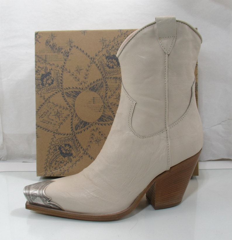 May include: Cream-colored cowboy boot with a silver-toned metal toe cap. The boot has a pointed toe, a stacked wooden heel, and a decorative stitch pattern. A cardboard box with a patterned design is in the background.