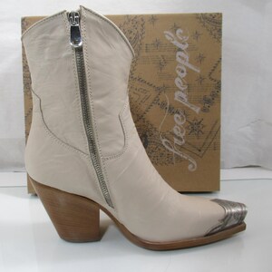 May include: Cream-colored leather cowboy boot with a silver zipper and toe cap. The boot has a wooden heel and is set against a brown box with the words "free people" printed on it.