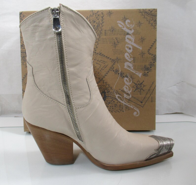 May include: Cream-colored leather cowboy boot with a silver zipper and toe cap. The boot has a wooden heel and is set against a brown box with the words "free people" printed on it.
