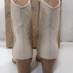 May include: A pair of beige leather ankle boots with wooden block heels. The boots have a side zipper and a pointed toe. The background features a cardboard box with a floral design.