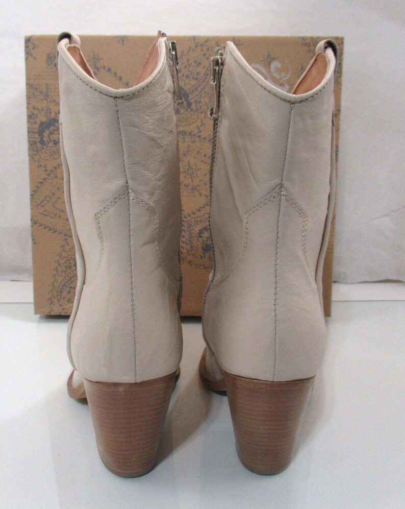 May include: A pair of beige leather ankle boots with wooden block heels. The boots have a side zipper and a pointed toe. The background features a cardboard box with a floral design.