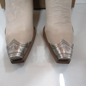 May include: Pair of off-white cowboy boots with silver-toned metal toe caps featuring an ornate design. The boots have a classic Western style with a pointed toe and a wooden sole.