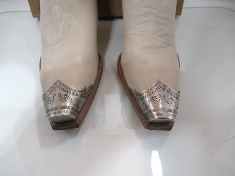 May include: Pair of off-white cowboy boots with silver-toned metal toe caps featuring an ornate design. The boots have a classic Western style with a pointed toe and a wooden sole.