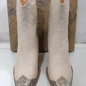 May include: A pair of off-white cowboy boots with silver-toned metal toe caps. The boots have a pointed toe and a mid-calf height. The background includes a brown box with a floral design.