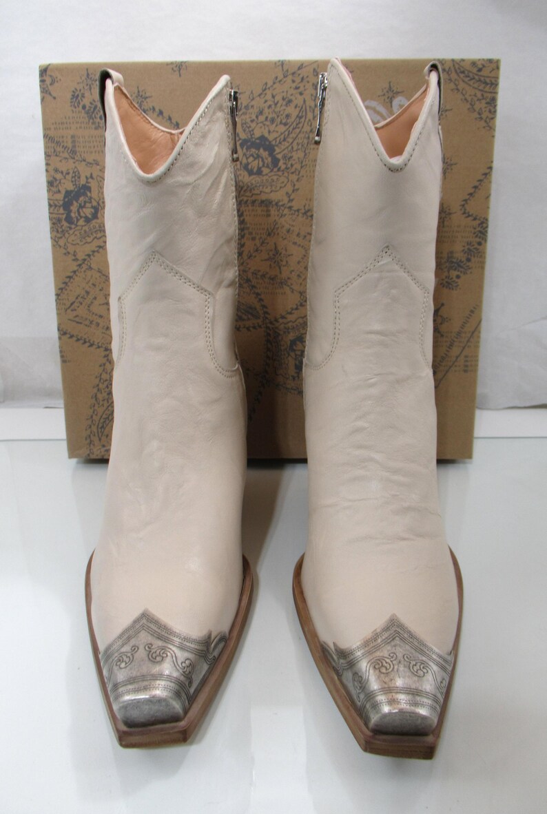 May include: A pair of off-white cowboy boots with silver-toned metal toe caps. The boots have a pointed toe and a mid-calf height. The background includes a brown box with a floral design.