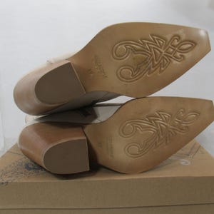 May include: A pair of beige leather ankle boots with a pointed toe and a stacked wooden heel. The soles feature a decorative design. The boots are displayed on a cardboard box.