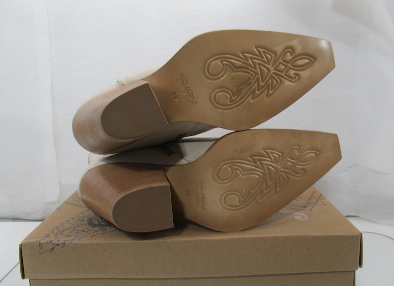 May include: A pair of beige leather ankle boots with a pointed toe and a stacked wooden heel. The soles feature a decorative design. The boots are displayed on a cardboard box.