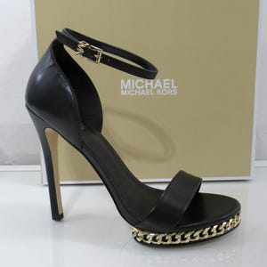 May include: Black leather high-heeled sandal with a gold chain detail around the platform sole. The sandal features a thin ankle strap with a buckle and the Michael Kors logo. The heel is a stiletto style.