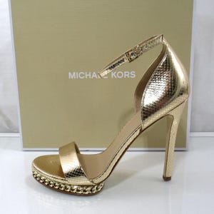 May include: A gold-colored high-heeled sandal with a stiletto heel and ankle strap. The sandal features a textured, snakeskin-like pattern and a platform sole with a chain detail. The brand name "MICHAEL KORS" is visible in the background.