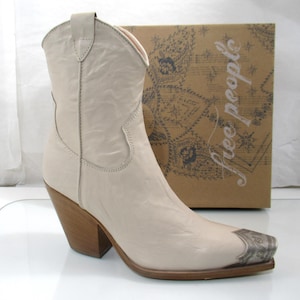 May include: Cream-colored cowboy boot with a pointed toe and a silver-toned metal toe cap. The boot has a stacked wooden heel and is set against a brown cardboard box with the words "free people" printed on it.