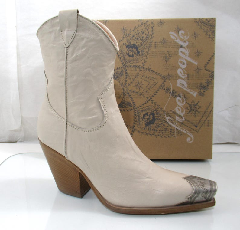 May include: Cream-colored cowboy boot with a pointed toe and a silver-toned metal toe cap. The boot has a stacked wooden heel and is set against a brown cardboard box with the words "free people" printed on it.