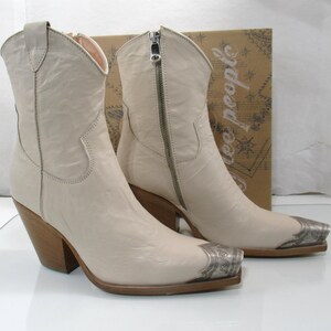 May include: A pair of off-white leather cowboy boots with silver-toned metal toe caps. The boots have a zipper closure, wooden heels, and a classic Western design. The boots are displayed in front of a cardboard box.