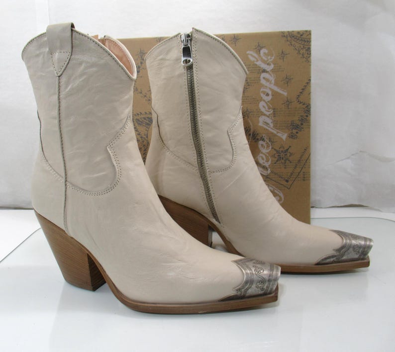 May include: A pair of off-white leather cowboy boots with silver-toned metal toe caps. The boots have a zipper closure, wooden heels, and a classic Western design. The boots are displayed in front of a cardboard box.