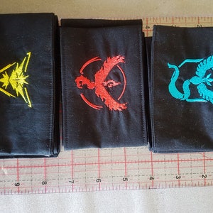 Può includere: Tre toppe di tessuto nero con disegni ricamati. La prima toppa ha un triangolo giallo con una stella e linee all'interno. La seconda toppa ha un uccello rosso con un cerchio intorno. La terza toppa ha un uccello blu con un serpente verde e un esagono intorno.