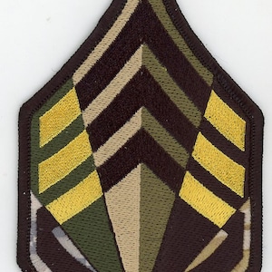 May include: A brown, tan, and green camouflage fabric military rank patch with three yellow stripes. The patch is shaped like a chevron.