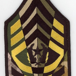 Puede incluir: Un parche de tela de camuflaje marrón, beige y verde con un diseño de chevron amarillo y una estrella negra en el centro. El parche tiene forma de triángulo con esquinas redondeadas.