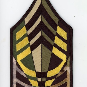 Puede incluir: Un parche de rango militar de camuflaje marrón, beige y verde con rayas de chevron amarillas. El parche tiene forma de chevron y tiene un borde negro.