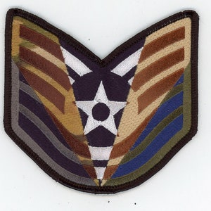 Puede incluir: Un parche de tela de camuflaje marrón, beige y verde con una estrella blanca y un centro azul oscuro. El parche tiene un borde marrón y tiene forma de chevron.