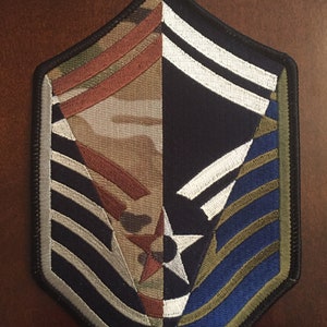 May include: A military patch with a black background and a star in the center. The patch has a camouflage pattern on the left side and a blue and green pattern on the right side. The patch has a silver border and is embroidered with white and silver stripes.