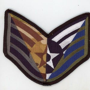 Puede incluir: Un parche de tela de camuflaje marrón, beige y blanco con una estrella blanca y un patrón de rayas azules y verdes. El parche está cosido sobre un fondo de tela marrón.