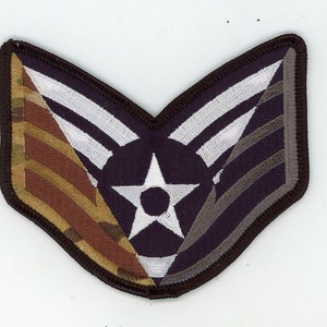 Puede incluir: Un parche de rango militar con una estrella de cinco puntas y dos rayas, en un patrón de camuflaje con colores marrón, verde y beige.