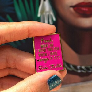 May include: A pink enamel pin with gold trim featuring a quote by Frida Kahlo: "Fest, what do I need you for when I have rings to fly?"