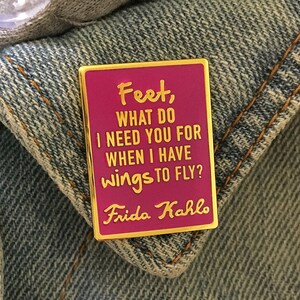May include: A pink enamel pin with a gold border featuring a quote by Frida Kahlo: "Feet, what do I need you for when I have wings to fly?"