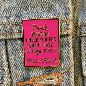 May include: Pink enamel pin with gold trim featuring a quote by Frida Kahlo: "Feet, what do I need you for when I have wings to fly?"