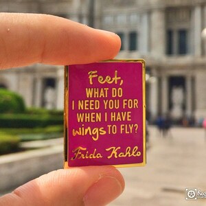 May include: A pink enamel pin with gold trim featuring a quote by Frida Kahlo: "Feet, what do I need you for when I have wings to fly?"
