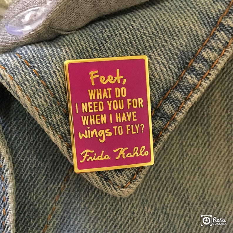 May include: A pink enamel pin with a gold border featuring a quote by Frida Kahlo: "Feet, what do I need you for when I have wings to fly?"