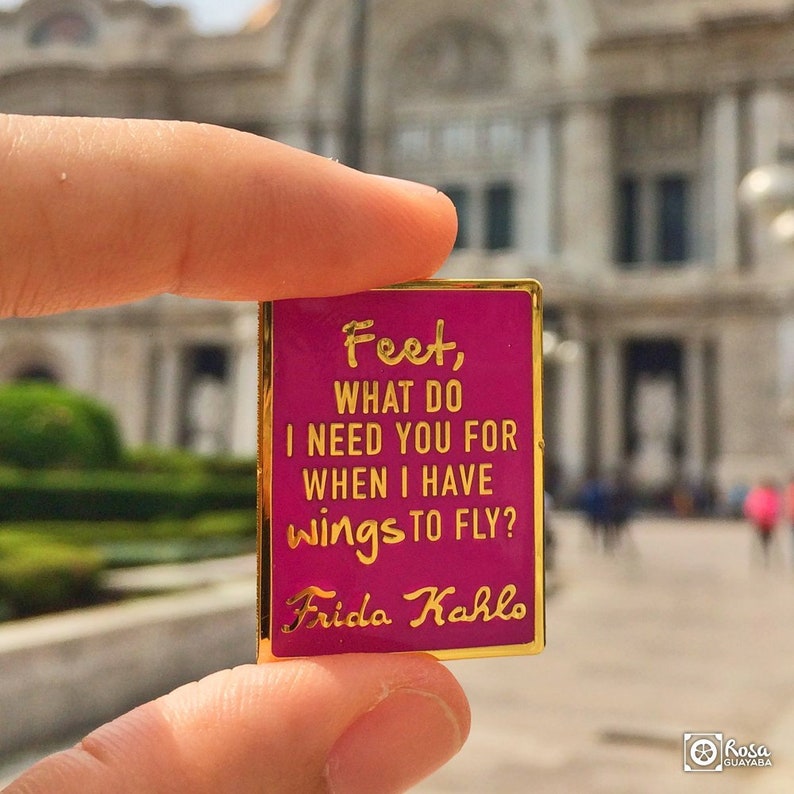 May include: A pink enamel pin with gold trim featuring a quote by Frida Kahlo: "Feet, what do I need you for when I have wings to fly?"