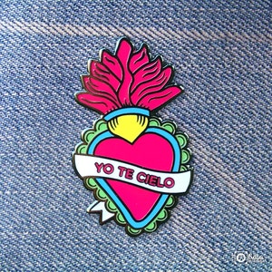 May include: A pink and blue enamel pin featuring a heart shape with a flame on top. The heart has a white banner with the text "YO TE CIELO" in black letters.