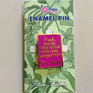 May include: Pink enamel pin with gold trim featuring a quote by Frida Kahlo: "Feet, what do I need you for when I have wings to fly?" The pin is displayed on a card with a green floral pattern.