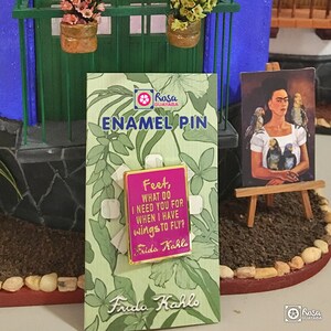 May include: A pink enamel pin with gold trim featuring a quote by Frida Kahlo: "Feet, what do I need you for when I have wings to fly?" The pin is displayed on a card with a green floral pattern and the text "Frida Kahlo" and "Enamel Pin".