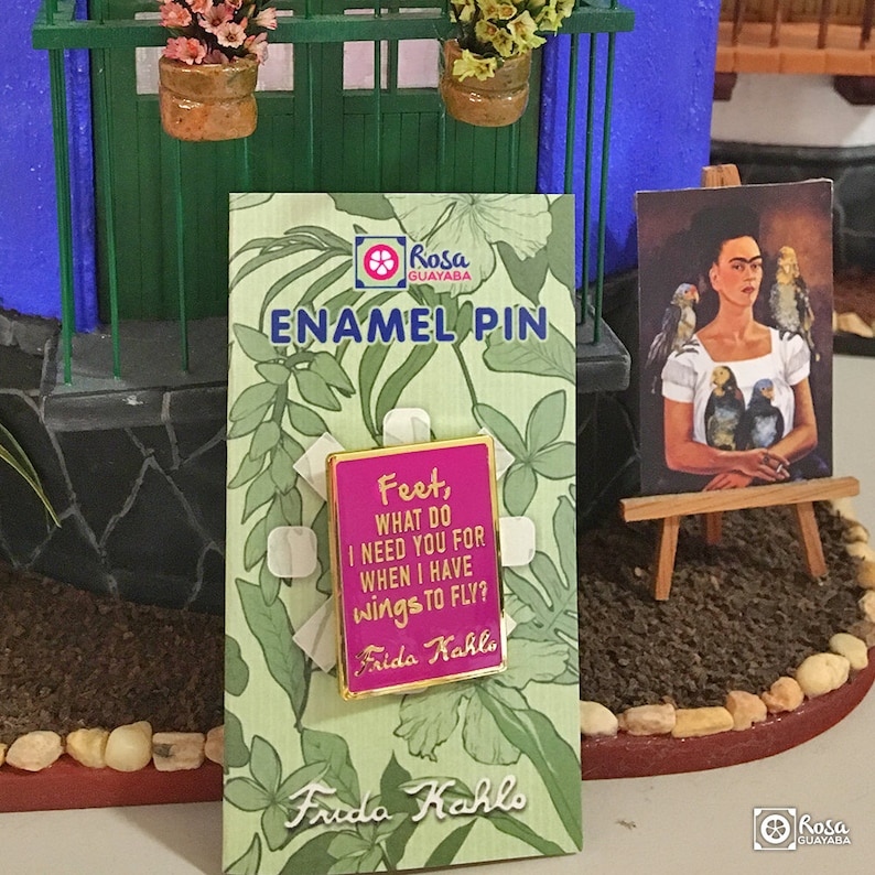 May include: A pink enamel pin with gold trim featuring a quote by Frida Kahlo: "Feet, what do I need you for when I have wings to fly?" The pin is displayed on a card with a green floral pattern and the text "Frida Kahlo" and "Enamel Pin".