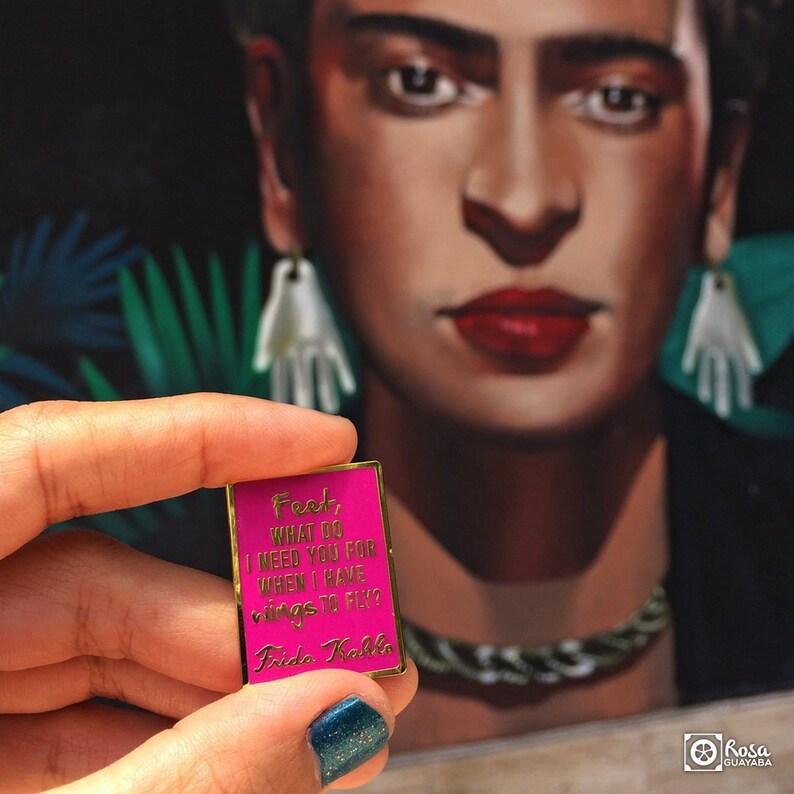 May include: A pink enamel pin with gold trim featuring a quote by Frida Kahlo: "Fest, what do I need you for when I have rings to fly?"
