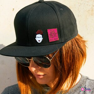 May include: Black baseball cap with a pink and black enamel pin featuring a portrait of Frida Kahlo and a quote: "Feet, what do I need you for when I have wings to fly." Frida Kahlo.