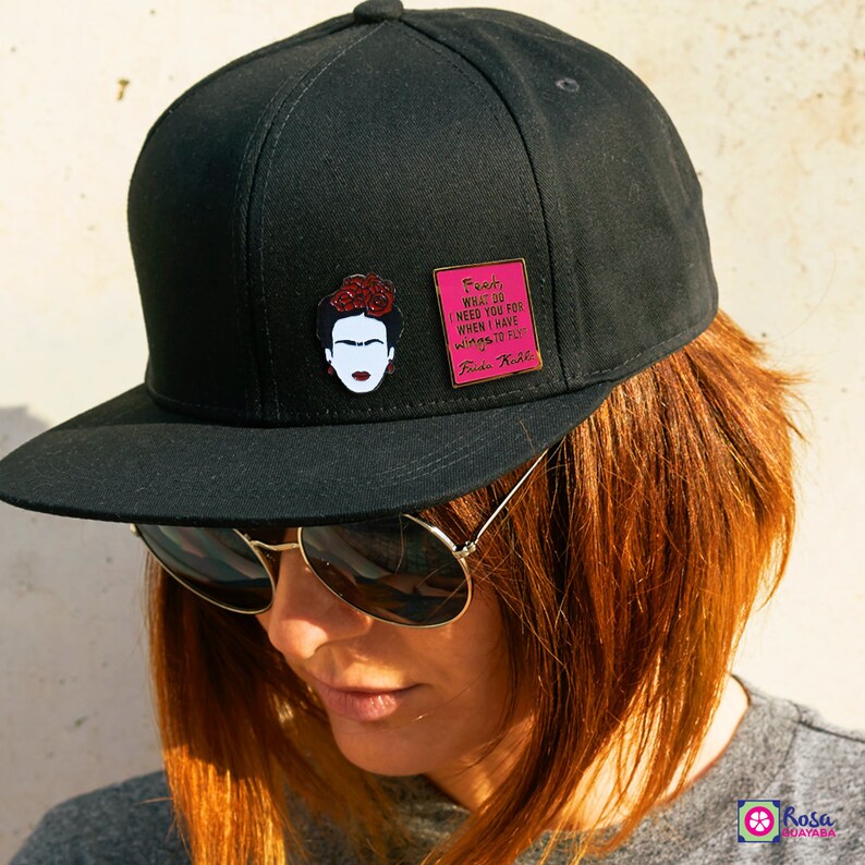 May include: Black baseball cap with a pink and black enamel pin featuring a portrait of Frida Kahlo and a quote: "Feet, what do I need you for when I have wings to fly." Frida Kahlo.