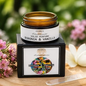 Könnte beinhalten: Ein kleiner bernsteinfarbener Glastiegel mit festem Parfüm, beschriftet mit Pearl's Natural Cosmetic Boronia & Vanilla, auf einer schwarzen Schachtel mit passendem Etikett und Blumengrafik. Ein durchsichtiger Kunststoffspatel liegt auf einer Holzoberfläche.