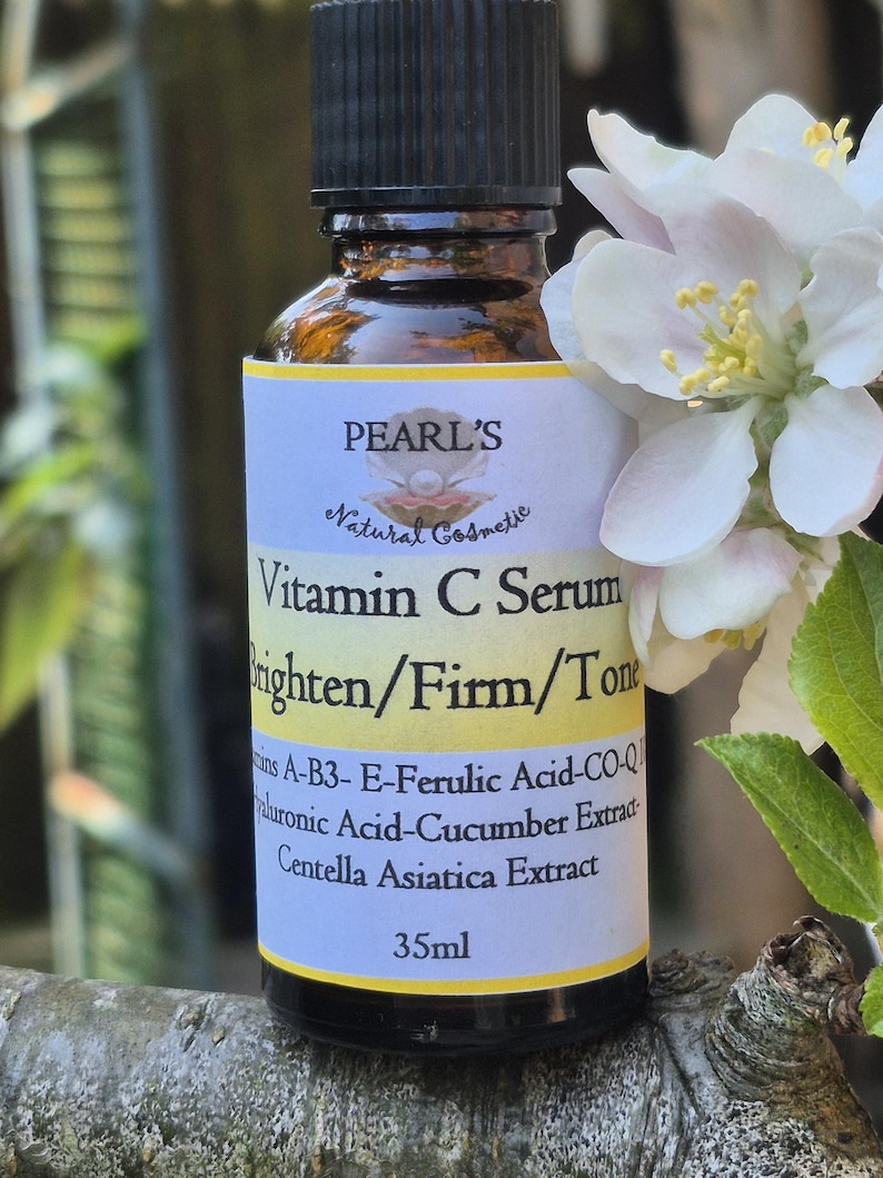 May include: A brown glass bottle of Pearl's Vitamin C Serum with a black cap. The label reads "Vitamin C Serum Brighten/Firm/Tone" and lists ingredients. A white flower with yellow stamens is in the background. The bottle contains 35ml.