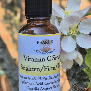 May include: A brown glass bottle of Pearl's Vitamin C Serum with a black cap. The label reads "Brighten/Firm/Tone" and lists ingredients. The bottle is 35ml. White flowers and green leaves are in the background.