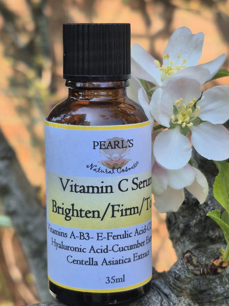 May include: A brown glass bottle of Pearl's Vitamin C Serum with a black cap. The label reads "Brighten/Firm/Tone" and lists ingredients. The bottle is 35ml. White flowers and green leaves are in the background.