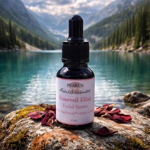 May include: A dark glass bottle of Pearl's Roseveil Elixir Facial Serum with a black dropper. The label is white with pink text. The bottle sits on a mossy rock with dried rose petals, with a lake and mountains in the background. The bottle contains 15ml.