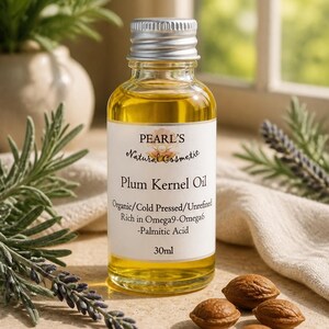 Può includere: Un flacone in vetro trasparente di olio di nocciolo di prugna Pearl's Natural Cosmetic con tappo argentato. L'etichetta riporta Organic/Cold Pressed/Unrefined, Rich in Omega9-Omega6-Palmitic Acid, 30ml. Quattro piccoli noccioli marroni sono davanti.