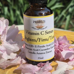 May include: A brown glass bottle of Pearl's Vitamin C Serum with a black cap, surrounded by pink flower petals. The label reads "Brighten/Firm/Tone" and lists ingredients. The bottle contains 35ml of serum.