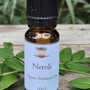 May include: A brown glass bottle of Neroli Organic Essential Oil, 10ml, with a black cap. The label is light blue with the brand name "Pearl's" and the product name. Green leaves are in the background.